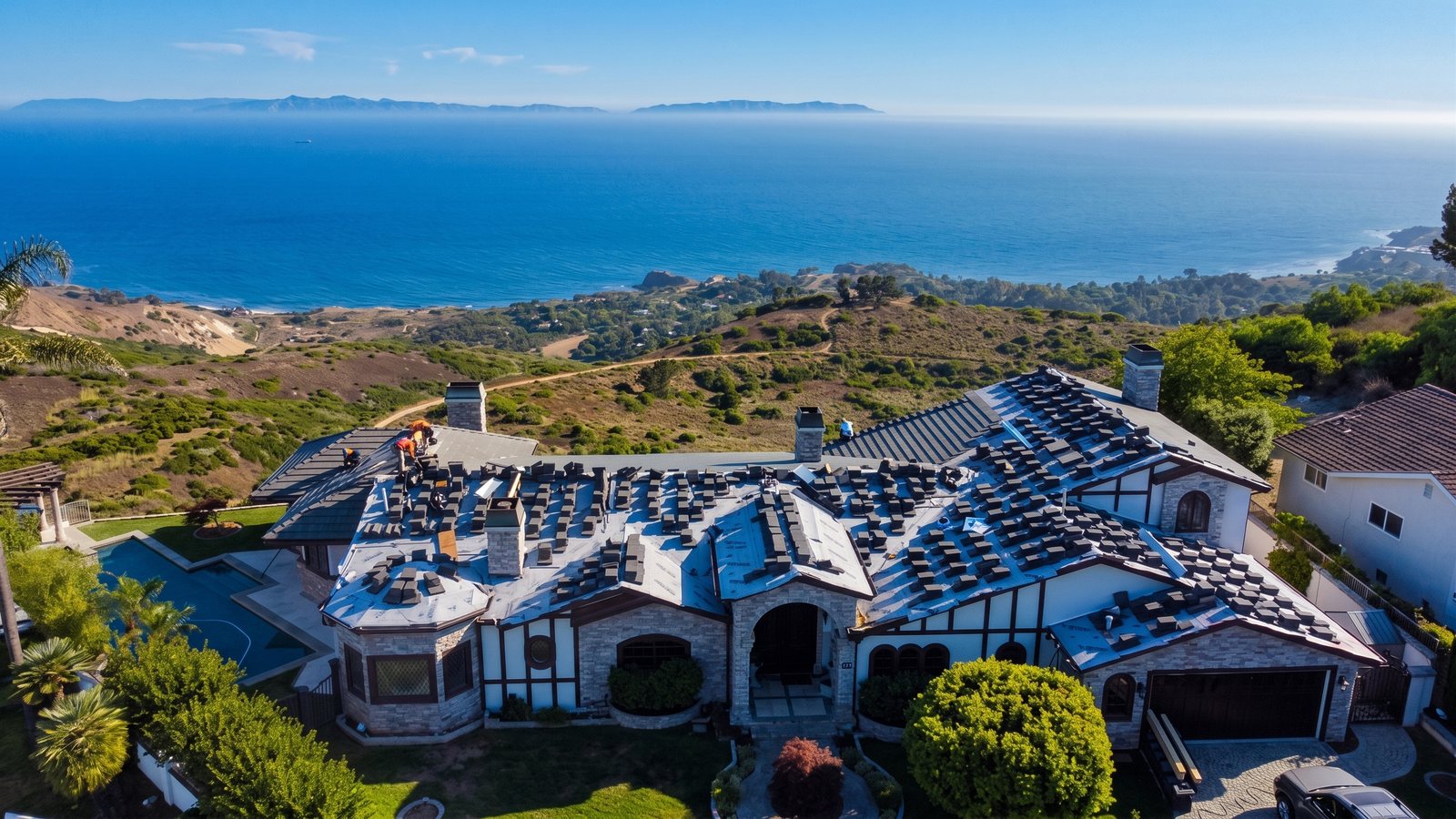 Drone view of a coastal estate mid re-roof, with new tile stacked across the roof and the ocean behind
